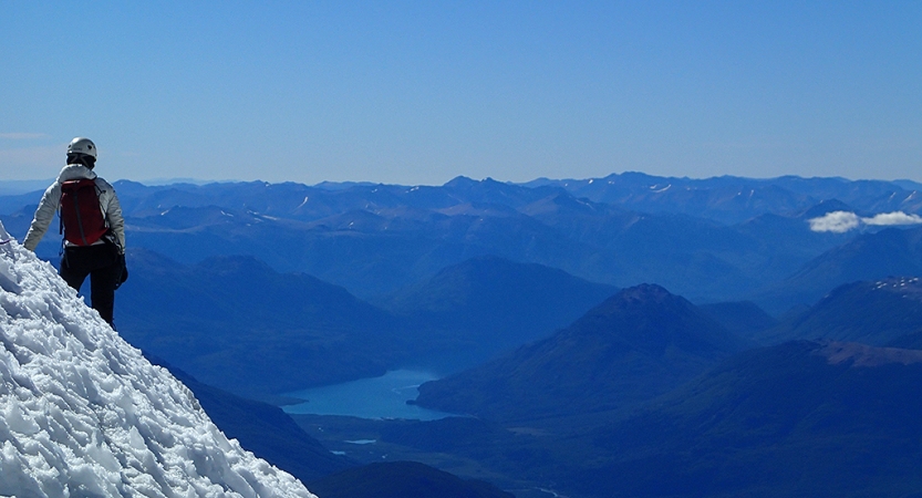 On the left side of the photo, a person wearing safety gear stands on a snowy incline, looking out over a vast mountainous landscape and a blue body of water. The mountains appear blue under the blue sky. 